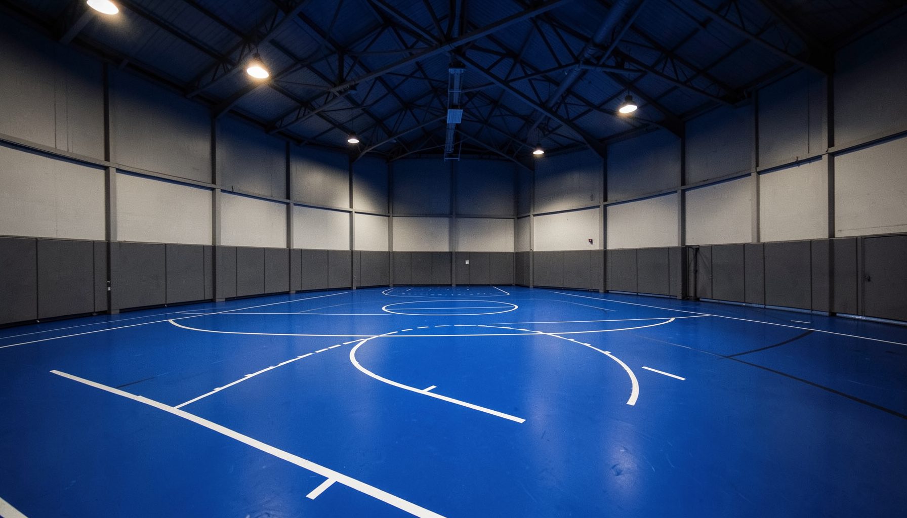 Wide-angle view of an indoor sports court with a vibrant blue polyurethane floor surface, white court markings, and warm overhead lighting